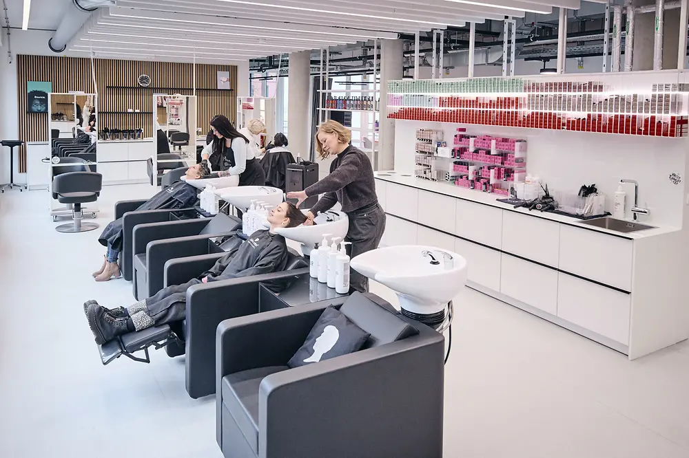 A photo of the modernly furnished Henkel test salon in Hamburg, showing three female clients seated in black chairs receiving hair treatments from hairdressers.