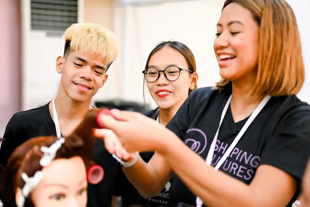 Two smiling young adults watch attentively as a Shaping Futures trainer styles the hair on a practice mannequin.